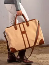 Bolso de viaje tipo duffel o carry-on, confeccionado en lona color beige claro con detalles, asas y correa de cuero marrón.