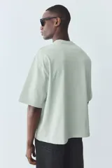 Remera verde claro de algodón pesado, corte oversized, con cuello redondo ribeteado, hombros caídos y bajo recto.