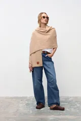 Chal de tejido de punto en color beige, con textura suave y diseño envolvente. Presenta una etiqueta de cuero sintético con logo grabado en la parte inferior.