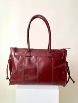 Bolso grande de cuero marrón con textura tipo cocodrilo, estructura rígida y asas para llevar al hombro. Tiene una tira con tachas y hebillas en cada extremo, también lleva lazos en los costados.
