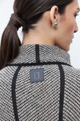 Capa de tejido de punto en color beige y negro con patrón de espiga, cuello alto y cierre de botón. Presenta vivos negros en contraste que delinean el diseño.