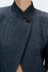 Blazer de manga corta confeccionado en lino, color azul marino, con cuello alto, cierre cruzado con un botón y pespuntes en contraste de color rojo. Cuenta con hombreras y bolsillos delanteros ribeteados.