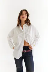 Camisa blanca de corte oversize, con cuello clásico, cierre frontal de botones y detalles de costuras trenzadas horizontales en el pecho. Presenta mangas largas con puños abotonados.
