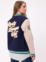 Campera estilo varsity con cuerpo en color marrón y mangas en tono beige. Presenta cierre frontal con botones a presión, cuello, puños y ruedo con franjas tejidas en verde y blanco. Incluye un parche con la letra R en el pecho y detalles bordados en las mangas.