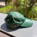 Gorra de cuero metalizado dorado, con ojales metálicos decorativos y etiqueta de marca aplicada en el lateral.