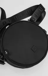 Bolso circular de estructura rígida, color negro, diseñado para el transporte de auriculares y accesorios. Cuenta con manija superior reforzada, correa de hombro ajustable con logo grabado, cierre perimetral y bolsillo trasero de acceso rápido.