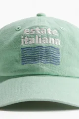 Gorra de sarga de algodón verde menta con visera curva y cierre ajustable con hebilla metálica en la parte trasera. Cuenta con bordado frontal con el texto "estate italiana" y diseño de ondas.