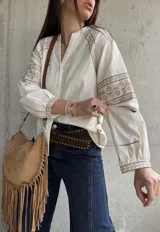 Camisa de inspiración boho de color crudo, con mangas largas abullonadas y puños decorados con detalles bordados geométricos en tonos tostados y dorados. Presenta cuello en V con cierre frontal de botones.