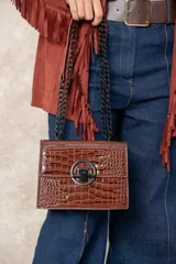 Cartera rígida de cuero sintético con acabado charol efecto cocodrilo, color chocolate. Presenta un herraje circular metálico negro con cierre central y cadena de eslabones negros como correa.