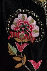 Chaqueta abierta de terciopelo negro con cuello redondo y mangas largas acampanadas. Presenta bordados florales en tonos rosados y verdes, con aplicaciones de abalorios y detalles dorados en el cuello y la parte frontal.