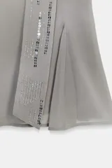 Top asimétrico color gris claro, con escote de un solo hombro. Presenta un tirante ancho decorado con lentejuelas plateadas que cae sobre el pecho y se anuda al cuello tipo halter.