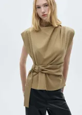 Blusa sin mangas color beige con cuello perkins y hombreras marcadas. Presenta un diseño recto con cinturón ajustable integrado en la cintura y cierre de cremallera en la parte posterior.