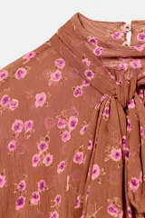 Camisa de estampado floral con cuello subido, lazo frontal y mangas largas con puños elásticos. Presenta un diseño con bajo asimétrico y cierre posterior con botones ocultos.