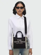 Cartera tipo tote de Marc Jacobs, modelo The Small Tote, confeccionada en lona negra con letras bordadas en color crema. Presenta doble asa superior, correa de hombro ajustable y extraíble, y detalles laterales en contraste.