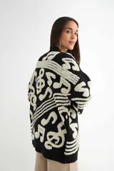 Saco tipo cardigan de punto, color negro con diseño de notas musicales y compases en color crema. Presenta cierre frontal con botones, cuello en V y puños acanalados.