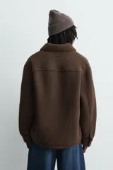 Sobrecamisa de corte relajado confeccionada en tejido con efecto borreguillo color marrón. Presenta cuello solapa, cierre frontal con botones a presión, mangas largas con puños abotonados y bolsillos frontales en la cadera.