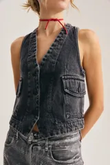 Chaleco de denim negro con efecto lavado, escote en V, cierre frontal de botones y bolsillos delanteros con solapa.
