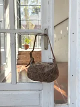 Bolso tipo hobo de cuero con acabado desgastado en tono visón. Presenta una correa ajustable con hebilla metálica y diseño de estructura redondeada.