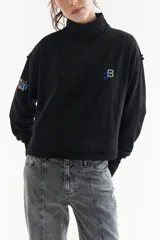 Buzo de algodón rústico con cuello polera, corte boxy y costuras externas visibles. Presenta mangas largas con puños con dedal y bordado decorativo en el pecho y la manga.
