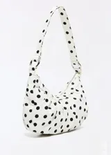 Cartera tipo hobo de diseño arqueado con estampado de lunares negros sobre fondo blanco. Cuenta con cierre superior de cremallera, tiras fruncidas decorativas en la base y una correa de hombro ajustable con argollas metálicas laterales.