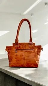 Cartera de cuero color marrón terra y dorado, con solapa de cuero texturizado, arte pintado a mano en la base, doble asa de hombro y herrajes metálicos.