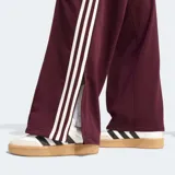 Pantalón deportivo Adidas Firebird de corte holgado, color bordó con las icónicas tres rayas laterales en blanco y logo del trébol bordado. Presenta cintura de tiro medio y diseño de pierna ancha.
