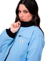 Campera tipo bomber de color azul claro, con cierre frontal, puños y cintura acanalados en color negro. Presenta un bolsillo pequeño con cierre en el pecho y el logo de la marca Caterpillar en una etiqueta rectangular. Cuenta con forro interior acolchado y diseño corto.
