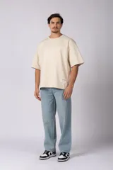Remera de corte oversize color beige, con cuello redondo y etiqueta decorativa pequeña en el ruedo inferior.