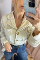 Campera tipo levita color crudo con textura efecto piel de durazno, cuello solapa, bolsillos frontales tipo cargo y cierre frontal con botones. Incluye un lazo ajustable en la cintura.