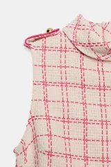 Vestido corto sin mangas de tejido texturizado con diseño de cuadros en tonos crudo y fucsia. Presenta cuello subido, trabillas decorativas con botones dorados en los hombros y cierre lateral con cremallera oculta.