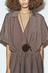 Collar de cordón fino negro con nudo, colgantes combinados de pieza metálica dorada, piedra ojo de tigre y cartera circular de piel 100% color marrón. Cierre con lazada y acabados con piezas metálicas doradas.