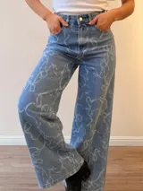 Jeans de corte wide leg y tiro alto, confeccionados en denim celeste con estampado de lazos en tono claro. Presentan cierre frontal con botón y cierre metálico, además de bolsillos funcionales.