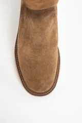 Bota de caña alta color camel, confeccionada en gamuza. Presenta una suela de goma gruesa y plana, y un diseño liso sin cierres visibles.