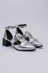 Sandalias plateadas con punta cerrada, tiras entrecruzadas y pulsera al tobillo con hebilla. Taco cuadrado negro.