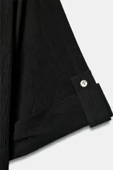 Camisa negra de textura arrugada, con cuello solapa, escote en V y cierre frontal de botones. Cuenta con mangas cortas terminadas en vuelta con trabilla y botón.