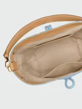 Bolso tipo bucket de denim azul claro con estampado floral blanco. Presenta detalles en cuero color tostado, asa de hombro fija y correa de tela ajustable extraíble. Incluye cierre frontal tipo toggle metálico dorado.