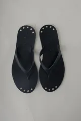 Sandalia plana tipo flip flop de cuero negro, con diseño minimalista y tira central de cuero. Presenta detalles de tachas metálicas en la base.