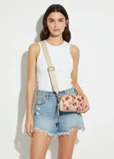 Mini cartera tipo bandolera con estampado de cerezas sobre fondo beige. Presenta una correa ancha ajustable de color crema, cierre superior con cremallera y herrajes metálicos dorados.