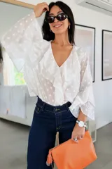Blusa blanca con estampado de lunares, confeccionada en tela semitransparente. Presenta escote en V con volados frontales y mangas largas acampanadas con volados en los puños.