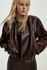 Chaqueta corta de piel color marrón, con cuello redondo, manga larga y cierre frontal con botones. Presenta un diseño con elástico en el ruedo y forro interior.