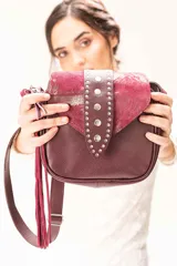 Cartera bandolera de cuero graneado y gamuza color bordó. Presenta una tapa frontal decorada con tachas metálicas, correa larga ajustable y un colgante lateral de flecos largos. Incluye bolsillo trasero con cierre.