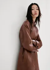 Chaqueta larga de efecto cuero color marrón, con cuello camisero, botonadura frontal oculta, bolsillos laterales y cinturón ajustable con hebilla en la cintura. Cuenta con trabillas en las muñecas y diseño amplio.