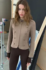 Blazer entallada color beige con rayas verticales finas, cuello redondo y manga larga. Presenta trabillas en los hombros y botones metálicos decorativos en el frente. Cierre frontal con cremallera y forro interior.