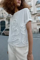 Camiseta de algodón de corte regular, con cuello redondo y manga corta. Presenta un detalle decorativo de flores con volumen confeccionadas en el mismo tejido sobre el frente.