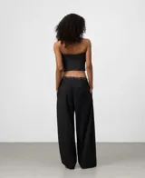 Pantalón negro de corte ancho y tiro alto con diseño asimétrico. Presenta un cruce frontal con cinturón de tela a cuadros grises y negros, que deja ver un forro o panel interior de la misma tela a cuadros.