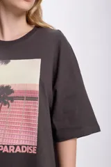 Remera de manga corta oversize color gris oscuro con estampa fotográfica de palmeras y edificio rosado.