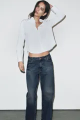 Camisa de popelín color blanco con cuello solapa redondeado y manga larga. Presenta un cierre frontal con botones ocultos y una tapeta decorada con perlas.