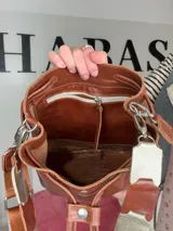 Bolso tipo bucket Chabas modelo Marin, confeccionado en cuero genuino de color marrón. Cuenta con diseño de cordón ajustable fruncido en la parte superior, manija corta decorativa de cuero con detalles metalizados y una correa larga intercambiable.