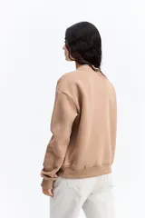 Buzo de algodón color beige con cuello redondo y estampa central rectangular en tonos rosa y dorado. Presenta un diseño de calce relajado con puños y ruedo acanalados.