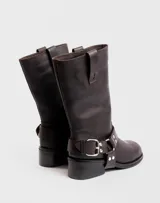 Bota de caña media confeccionada en cuero vacuno color marrón oscuro, con diseño de estilo biker. Presenta una tira decorativa con hebilla metálica y estribo desmontable en la zona del tobillo, taco cuadrado de madera y suela de caucho.